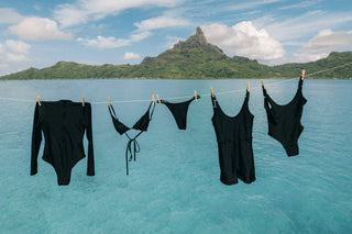 Black swimwear items hanging on a clothesline over clear turquoise water, with a tropical island and mountain in the background.